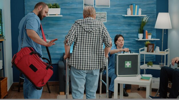 Caregiver supporting senior patient using walking frame during in-home medical visit with equipment nearby.