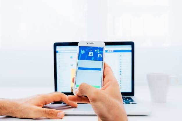 How to Measure the Success of Your Facebook Ads Campaigns 2 Person holding smartphone with Facebook login screen in front of laptop, managing social media account.